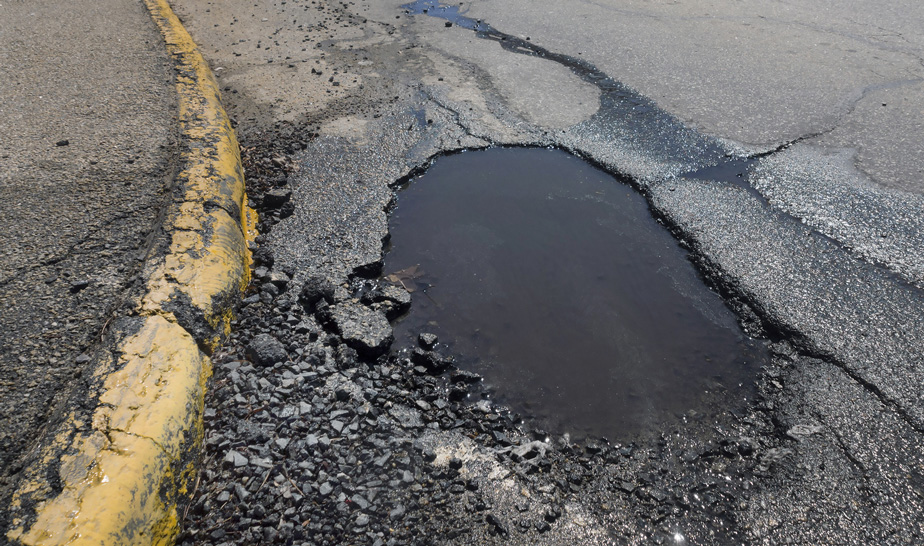A broken asphalt and pothole filled with rainwater near a yellow-painted curb.