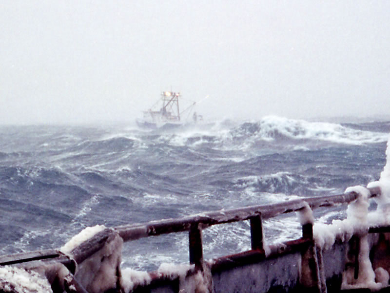 The view from the deck of a ship, over the ice-covered railing. A commercial ship can be seen navigating rough seas in the distance.
